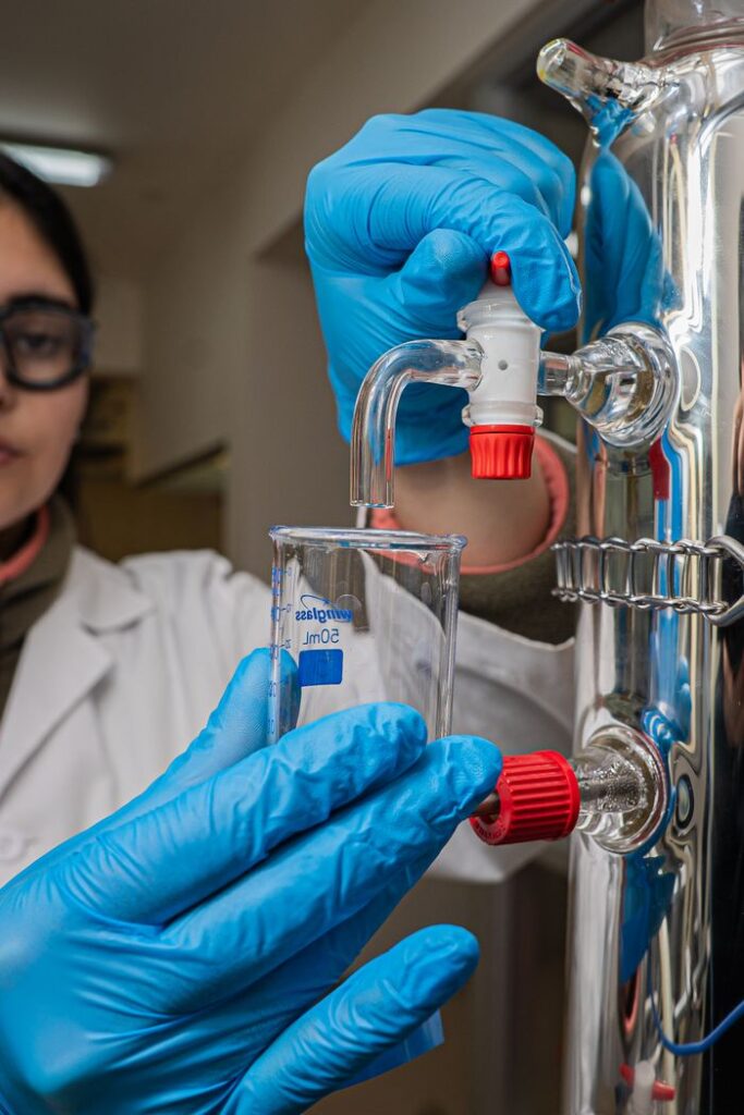 A scientist wearing gloves and goggles adjusts a distillation apparatus in a chemistry lab.